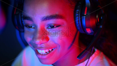 Black teen girl in headset playing video games in video game club with blue and red illumination, laughing and talking in voice chat. Slow motion - Starpik