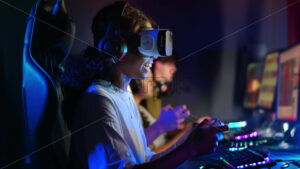 Black teen smiling girl in VR headset playing video games in video game club with blue illumination using a gamepad, virtual reality - Starpik Stock