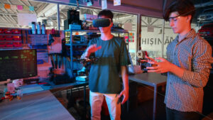 Teens doing experiments in robotics in a laboratory. Boy in protective glasses and guy in VR headset controlling a flying drone using controller and hand. Red and blue illumination - Starpik