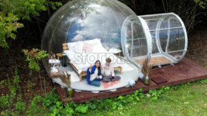 A couple sitting inside a transparent bubble tent at glamping. Sitting under a blanket, holding cups and talking. Lush forest on the background - Starpik Stock