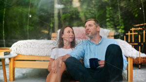 A talking couple sitting inside transparent bubble tent at glamping with cups. Lush forest on the background - Starpik Stock