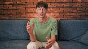 A young man blogger influencer talking and looking into the camera while sitting on a sofa - Starpik Stock