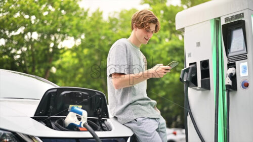 A young man using smartphone at a car charging station with charging electric car nearby. Chisinau, Moldova. Slow motion - Starpik Stock