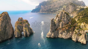 Aerial drone cinematic view of the Tyrrhenian sea coast of Capri, Italy. Rocky cliffs, blue water, floating boats, greenery, town in the distance - Starpik