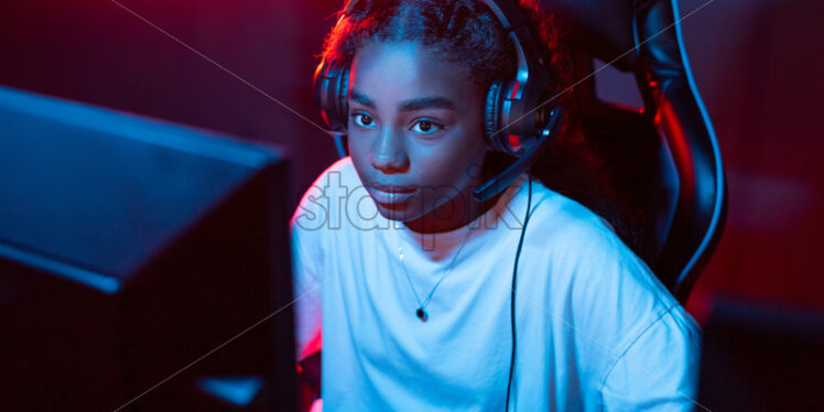 Black teen girl in headset playing video games in video game club with blue and red illumination - Starpik