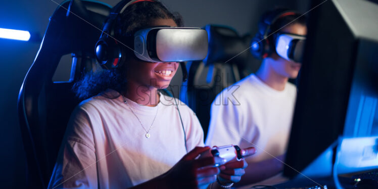 Black teen smiling girl in VR headset playing video games in video game club with blue illumination using a gamepad - Starpik