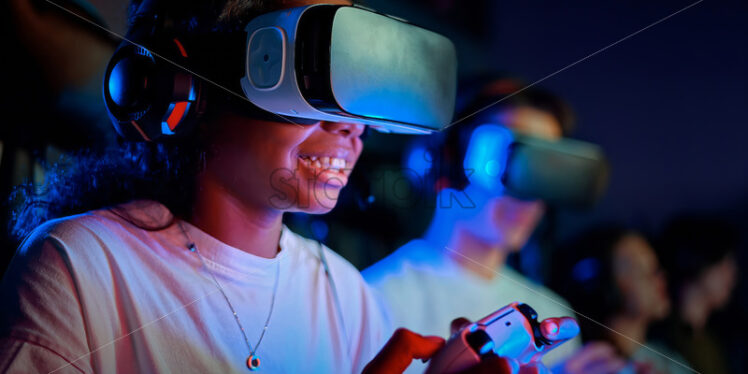 Black teen smiling girl in VR headset playing video games using gamepad in video game club - Starpik