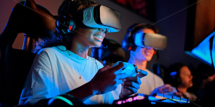 Black teen smiling girl in VR headset playing video games using gamepad in video game club with blue illumination - Starpik