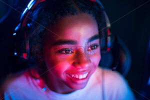 Black teen smiling girl in headset playing video games in video game club with blue and red illumination - Starpik