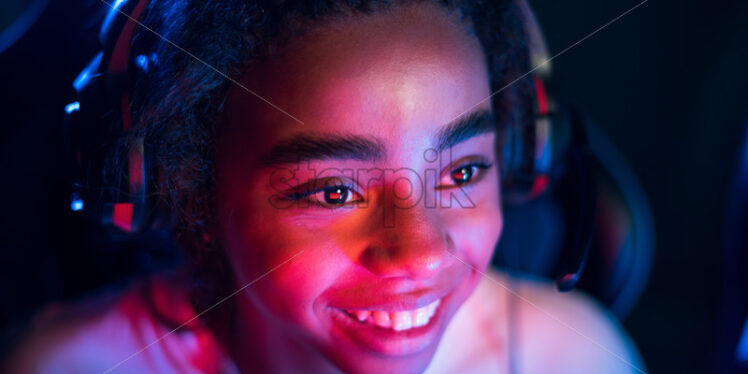 Black teen smiling girl in headset playing video games in video game club with blue and red illumination - Starpik