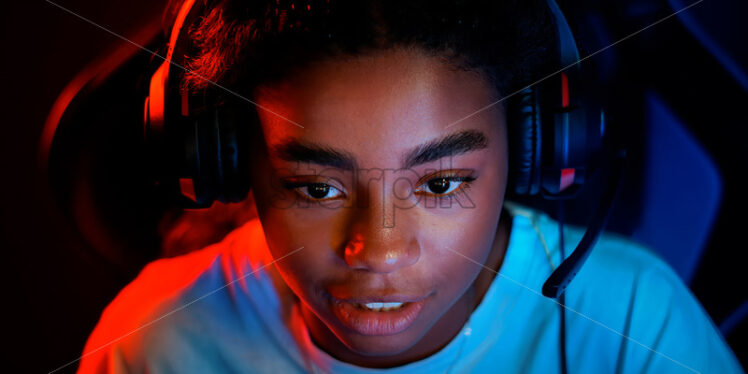 Black teen smiling girl in headset playing video games in video game club with red illumination - Starpik