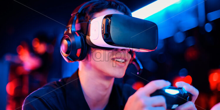 Close view of a smiling teen playing a game console in VR headset and headphones using gamepad. Blue and red illumination - Starpik Stock