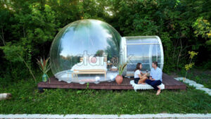 Drone view of a happy couple drinking wine near transparent bubble tent at glamping. Nature around - Starpik Stock