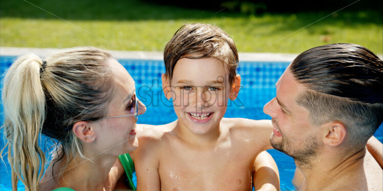 Mother with father and son resting and swimming in a pool in summer, happy family kissing - Starpik Stock