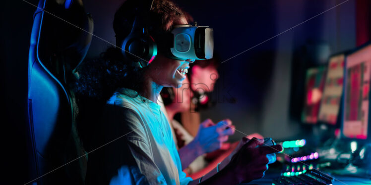 Premiu stock photo – Black teen smiling girl in VR headset playing video games using gamepad in video game club with blue illumination - Starpik