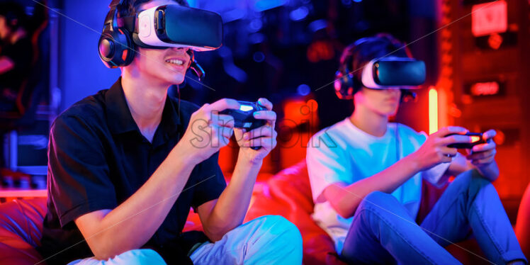 Premium stock photo –Two teen friends are playing a game console in VR headset and headphones using gamepads and smiling while sitting on bean bags. Blue and red illumination - Starpik