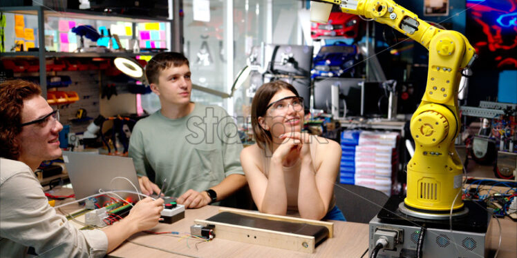Three young happy engineers fixing an yellow robotic arm in the workshop, computer programming - Starpik Stock