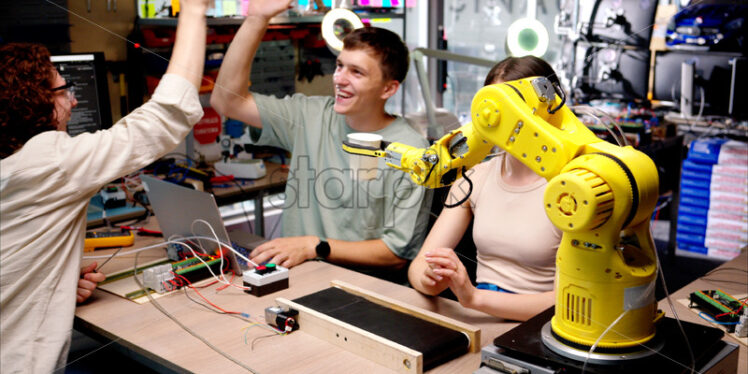 Three young happy engineers fixing an yellow robotic arm in the workshop, computer programming, celebrating success - Starpik Stock