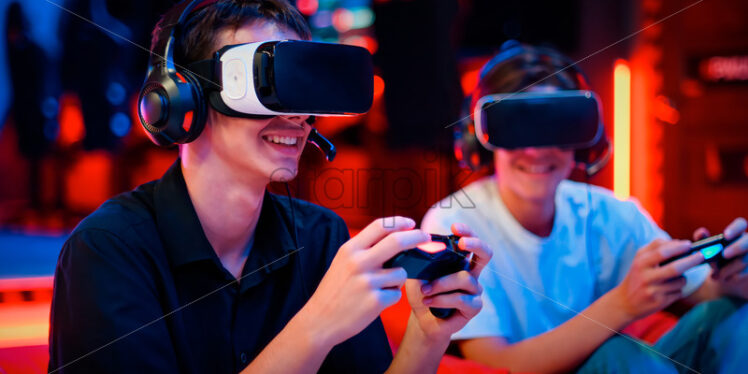 Two teen friends are playing a game console in VR headset and headphones using gamepads and smiling while sitting on bean bags. Blue and red illumination - Starpik Stock