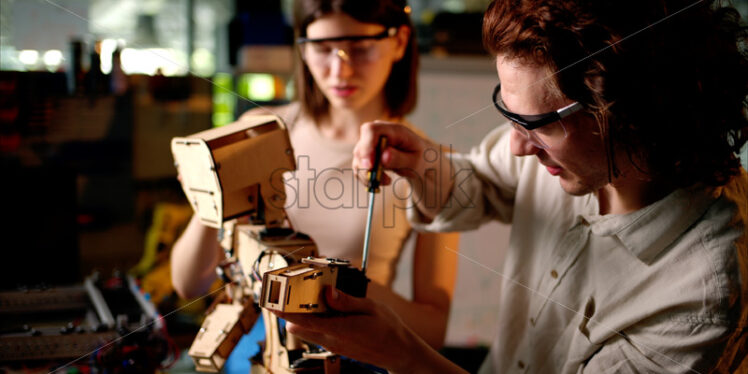Two young engineers fixing a mechanical robot in the workshop, wearing industrial glasses - Starpik Stock