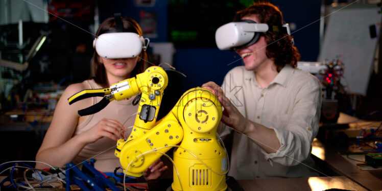 Two young engineers fixing a yellow robot arm in the workshop, wearing VR virtual reality headset - Starpik Stock
