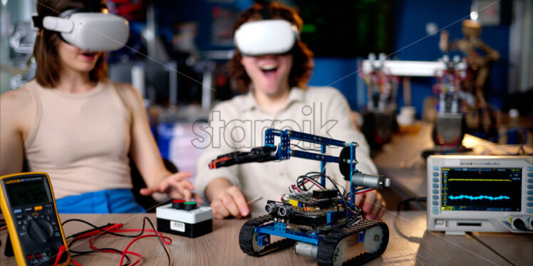 Two young happy engineers fixing a mechanical robot car in the workshop, using VR virtual reality headsets - Starpik Stock