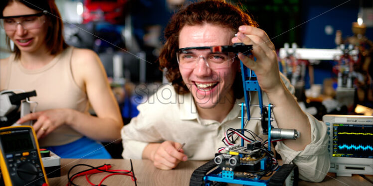 Two young happy engineers fixing a mechanical robot car in the workshop, using VR virtual reality headsets - Starpik Stock