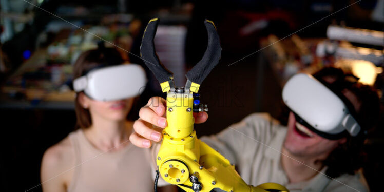 Two young happy engineers fixing an yellow robotic arm in the workshop, computer programming, celebrating success - Starpik Stock