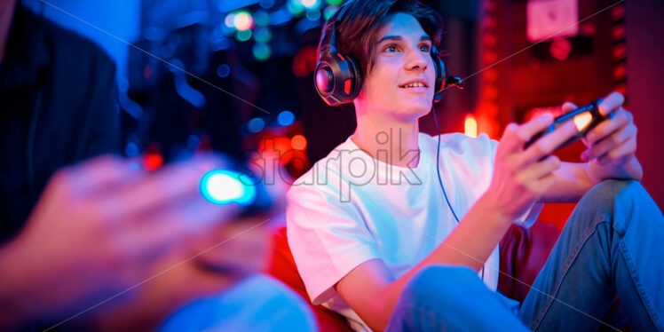 View of a smiling teen playing a game console in headphones using gamepad while sitting on bean bag. Blue and red illumination - Starpik Stock