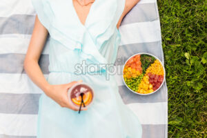 Woman laying on a blanket in the park holding a drink and poke bowl near her - Starpik Stock