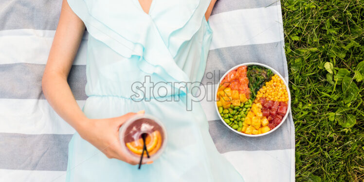 Woman laying on a blanket in the park holding a drink and poke bowl near her - Starpik Stock