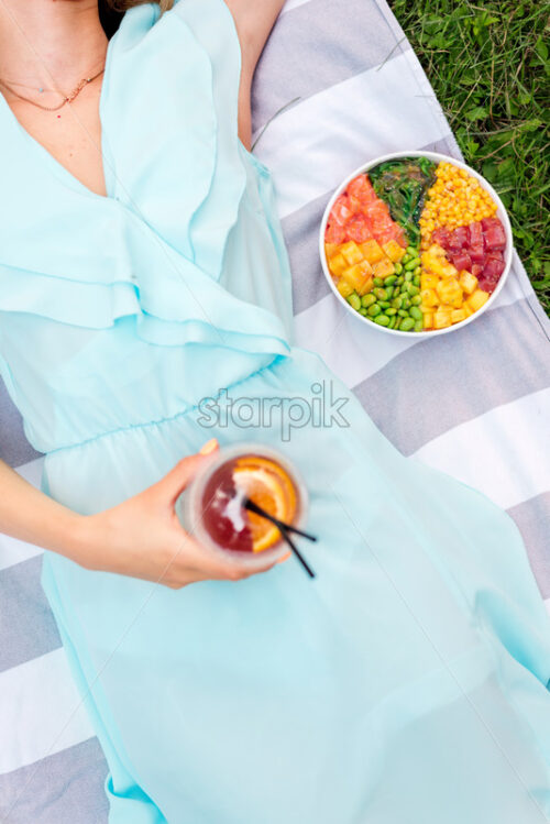 Woman laying on a blanket in the park holding a drink and poke bowl near her - Starpik Stock