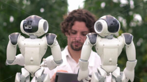 VIDEO Laboratory technician in a white coat holding a tablet and analysing two humanoid robots in a greenhouse farm - Starpik