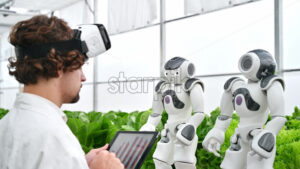 VIDEO Laboratory technician in a white coat wearing virtual reality headset analysing graphs on a tablet while interacting with two humanoid robots in a greenhouse farm - Starpik