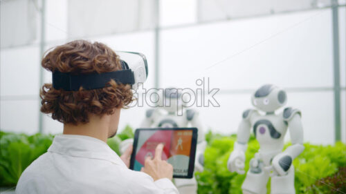 VIDEO Laboratory technician in a white coat wearing virtual reality headset analysing graphs on a tablet while interacting with two humanoid robots in a greenhouse farm - Starpik