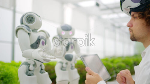 VIDEO Laboratory technician in a white coat wearing virtual reality headset analysing graphs on a tablet while interacting with two humanoid robots in a greenhouse farm - Starpik