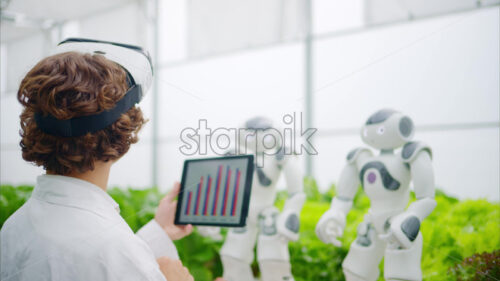 VIDEO Laboratory technician in a white coat wearing virtual reality headset analysing graphs on a tablet while interacting with two humanoid robots in a greenhouse farm - Starpik