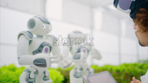 VIDEO Laboratory technician in a white coat wearing virtual reality headset analysing graphs on a tablet while interacting with two humanoid robots in a greenhouse farm - Starpik