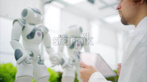 VIDEO Laboratory technician in a white coat wearing virtual reality headset analysing graphs on a tablet while interacting with two humanoid robots in a greenhouse farm - Starpik