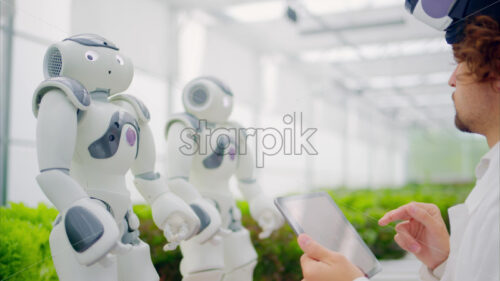 VIDEO Laboratory technician in a white coat wearing virtual reality headset analysing graphs on a tablet while interacting with two humanoid robots in a greenhouse farm - Starpik