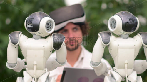 VIDEO Laboratory technician in a white coat with virtual reality headset holding a tablet while analysing two humanoid robots in a greenhouse farm - Starpik