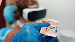 Video Dentist wearing a protective mask, goggles, and gloves showing a teeth mock up while the patient is wearing a Virtual Reality headset in dental cabinet - Starpik