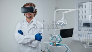 Video Dentist wearing a scrub and a Virtual Reality headset while standing and smiling in a dental cabinet, in front of a window - Starpik