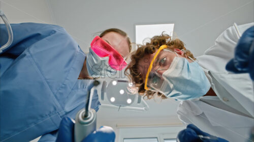 Video Low angle view of two dentists wearing protective masks, goggles, and gloves, holding dental instruments under bright procedural lights - Starpik