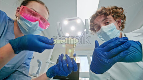 Video Low angle view of two dentists wearing protective masks, goggles, and gloves, showing a big tooth and brush figurine under bright procedural lights - Starpik