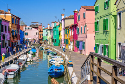 BURANO, ITALY – APRIL 03, 2017: Daylight view to a street with colorful buildings and people walking on sidewalk. Parked boats in canal and image reflecting on water - Starpik