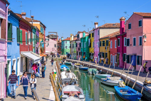 BURANO, ITALY – APRIL 03, 2017: Daylight view to a street with colorful buildings and people walking on sidewalk. Parked boats in canal and image reflecting on water. Bright blue sky - Starpik