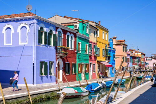 BURANO, ITALY – APRIL 03, 2017: Daylight view to a street with colorful buildings and people walking on sidewalk. Parked boats in canal and image reflecting on water. Bright blue sky - Starpik