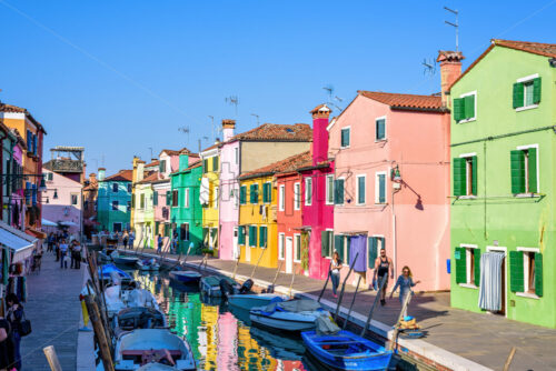 BURANO, ITALY – APRIL 03, 2017: Daylight view to a street with colorful buildings and people walking on sidewalk. Parked boats in canal and image reflecting on water. Bright blue sky - Starpik