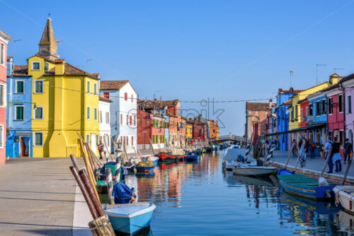 BURANO, ITALY – APRIL 03, 2017: Daylight view to a street with colorful buildings and people walking on sidewalk. Parked boats in canal and image reflecting on water. Bright blue sky - Starpik